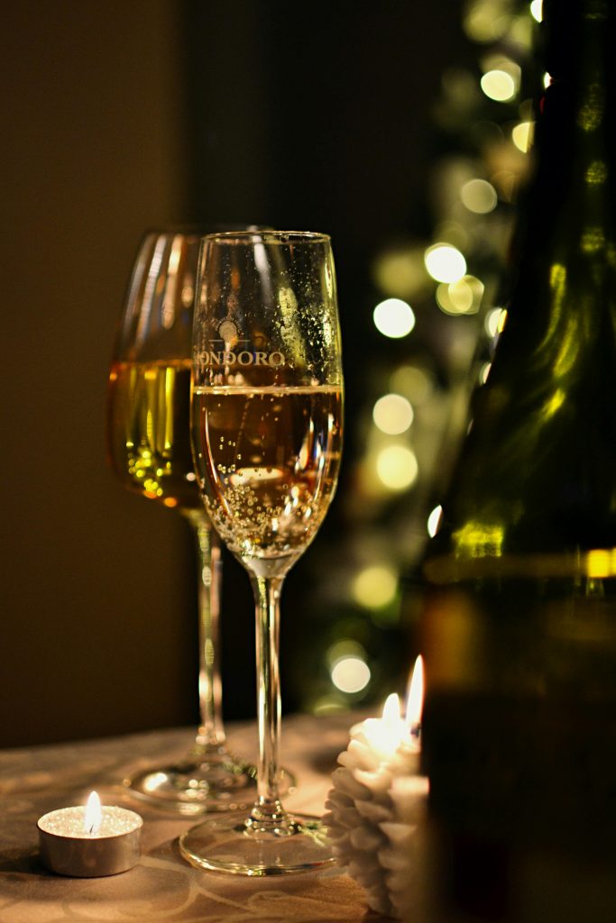 Zwei Gläser mit prickelndem Sekt und Wein vor einem Weihnachtsbaum, inmitten von Kerzen - so kann man sich auf ein gemütliches Silvester in Loungewear einstimmen.